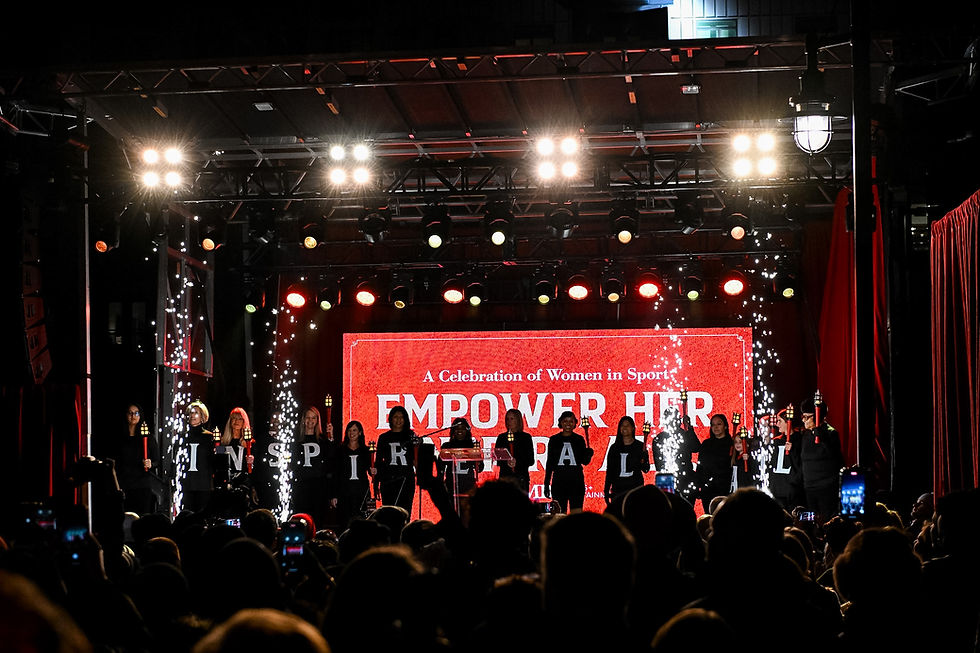 Women on stage hold letters forming "INSPIRE ALL" amid sparklers. Red backdrop reads "EMPOWER HER" in a sports celebration.