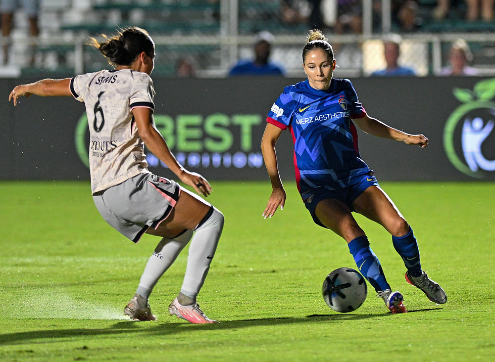 Two women soccer players contest the ball on a lit field at night, one in blue MERZ AESTHETICS kit and one in white.