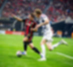 Two soccer players compete for the ball on a green field. One wears a red and black uniform, the other white and blue. Crowd in background.