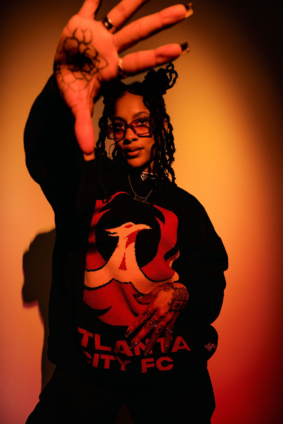 Stylish woman with dreadlocks and glasses poses under warm orange light, hand raised; black shirt reads Atlanta City FC.
