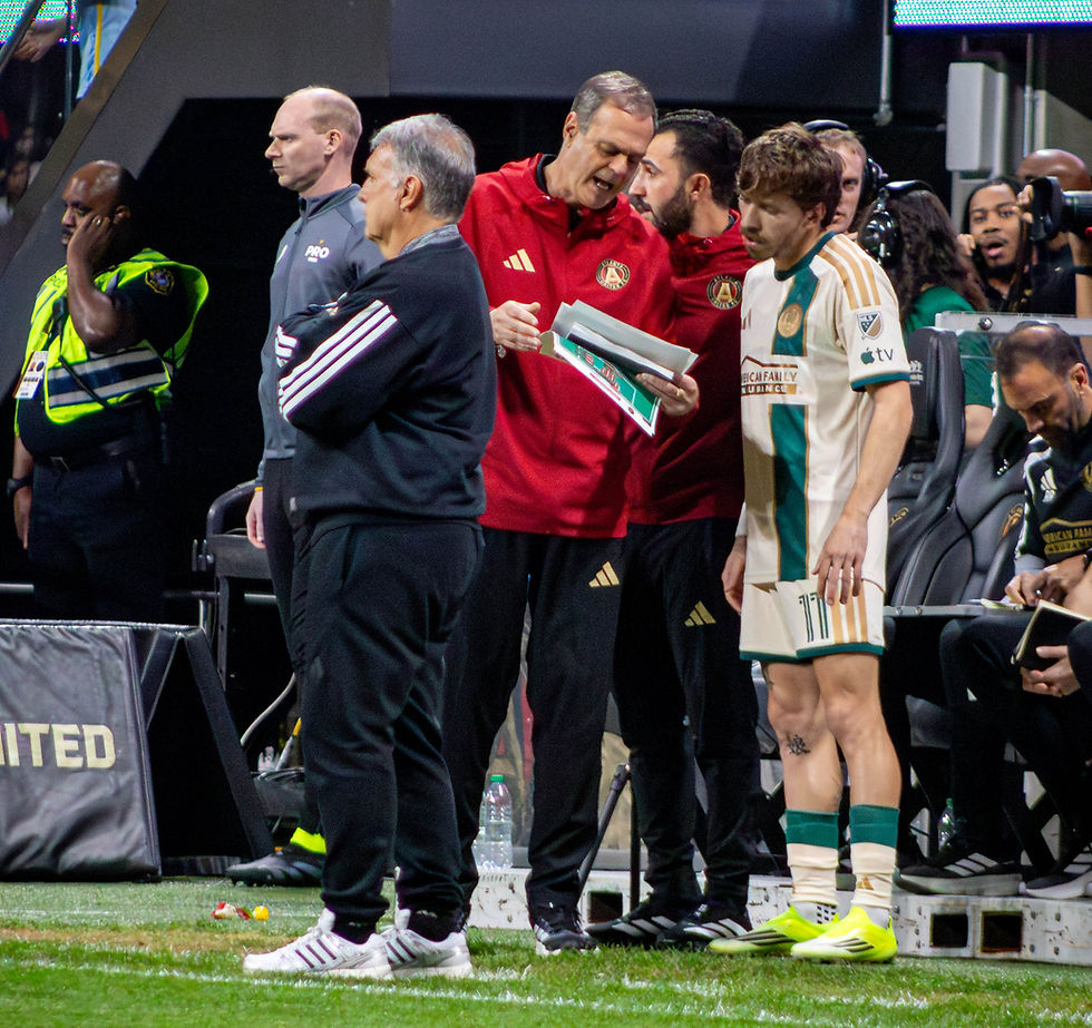 Soccer coach in red gives instructions from a clipboard to a player in a white and green jersey on the sidelines. Staff and security nearby.