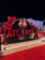 Red and black display reading "ATLANTA 2028" with NWSL logo, surrounded by red balloons on a red carpet. Night setting, festive mood.