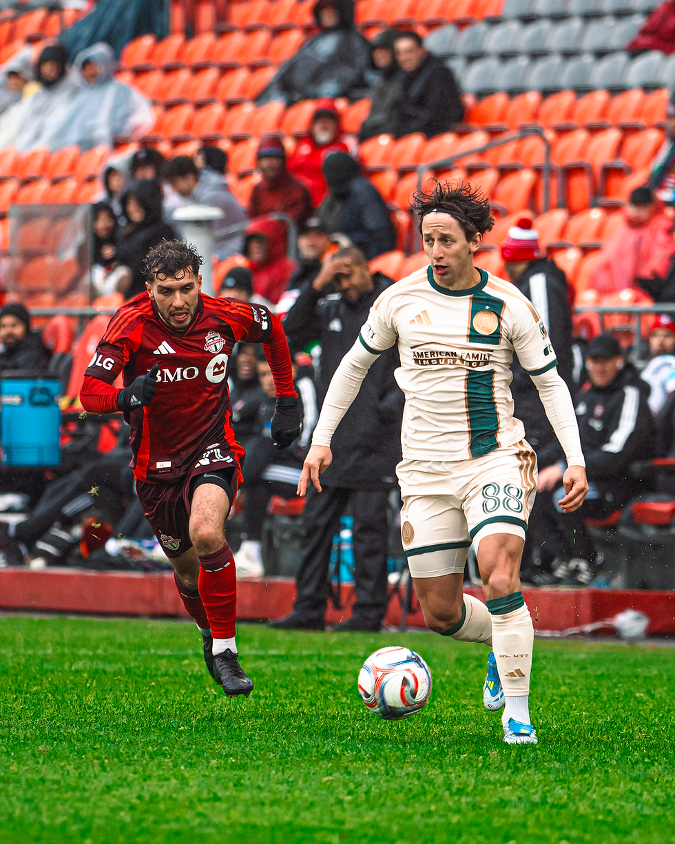 Two soccer players in action on a grassy field; one in red, one in white with green. Crowd in orange seats in the rainy background.