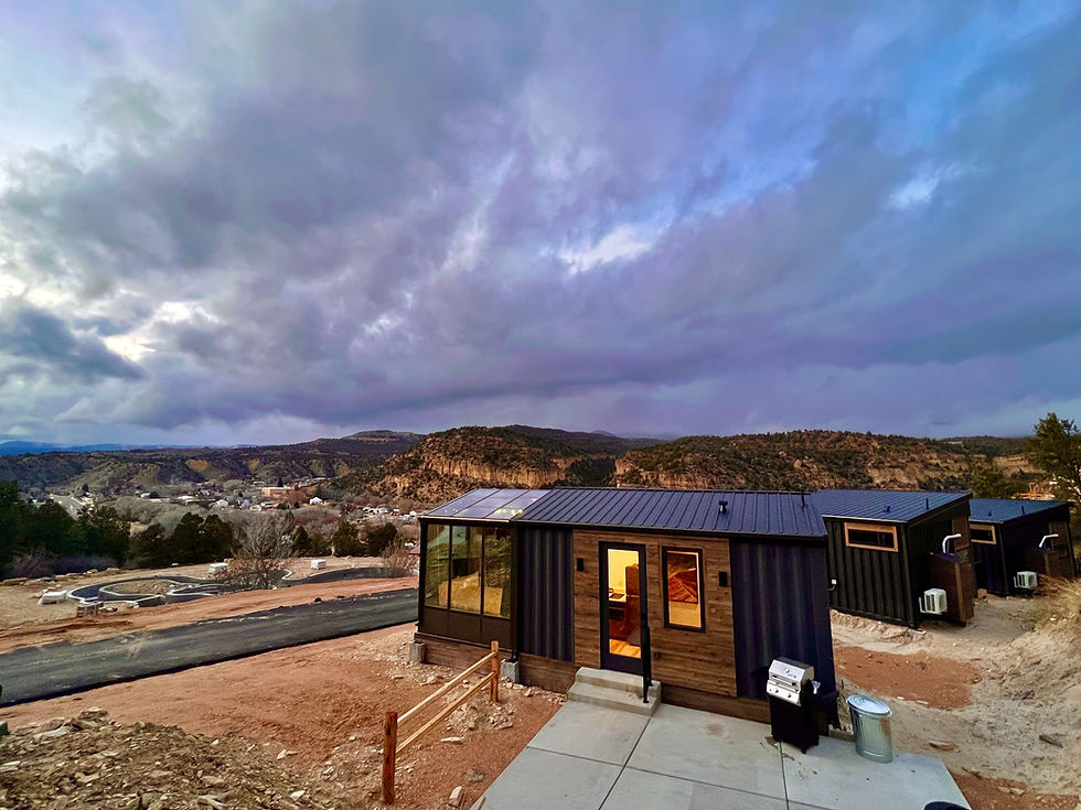 Modern container tiny homes sit on a hillside overlooking a mountainous valley under a dramatic, cloudy storm sky. The main cabin features wood paneling, an attached glass sunroom, and warm indoor lighting, with a barbecue grill on the patio outside.