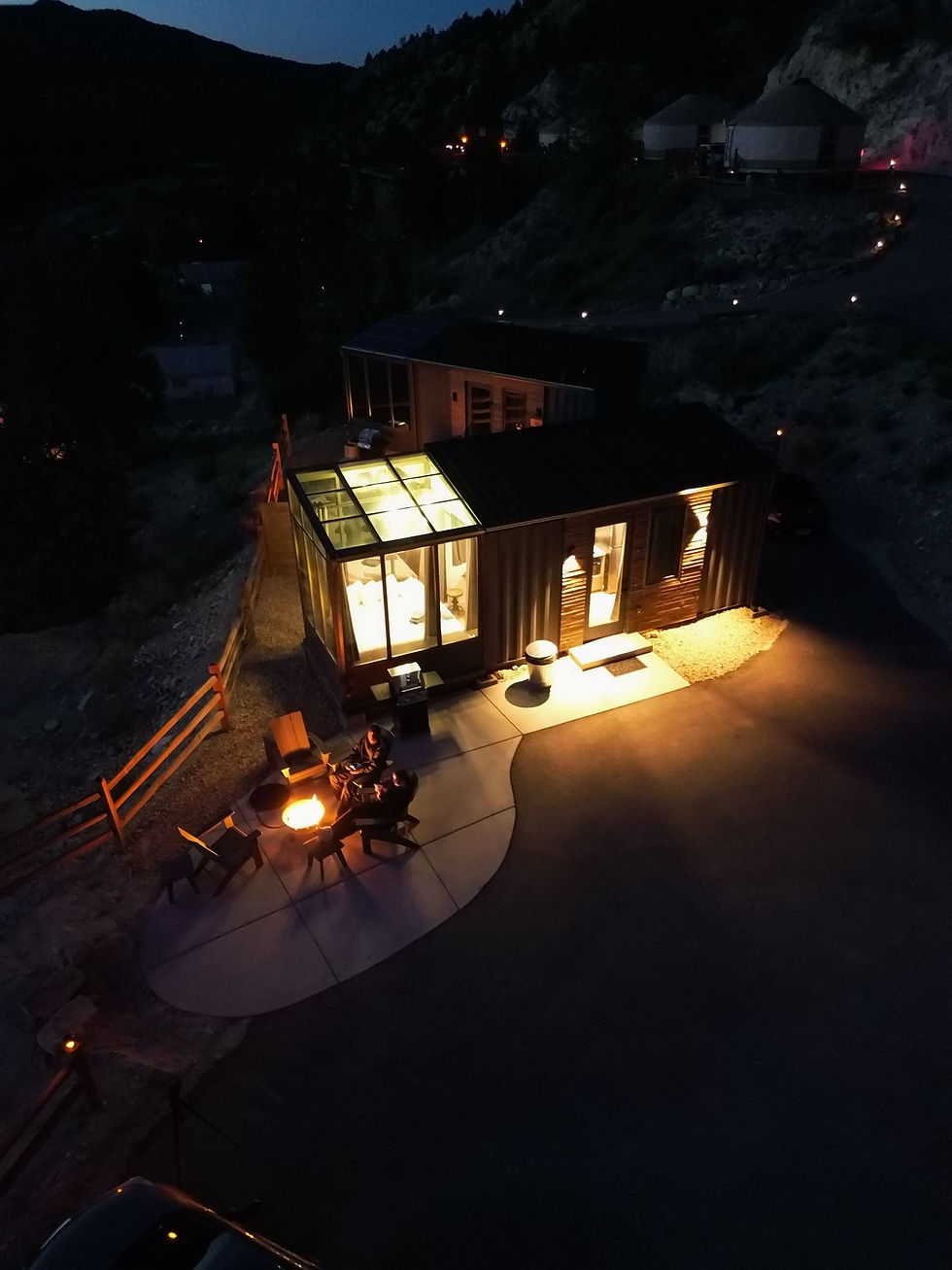 An aerial, nighttime view of a modern container tiny home with a brightly lit glass sunroom and a patio fire pit.