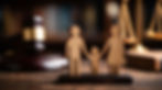 Wooden family figurines holding hands, with a gavel and scales of justice in the background.