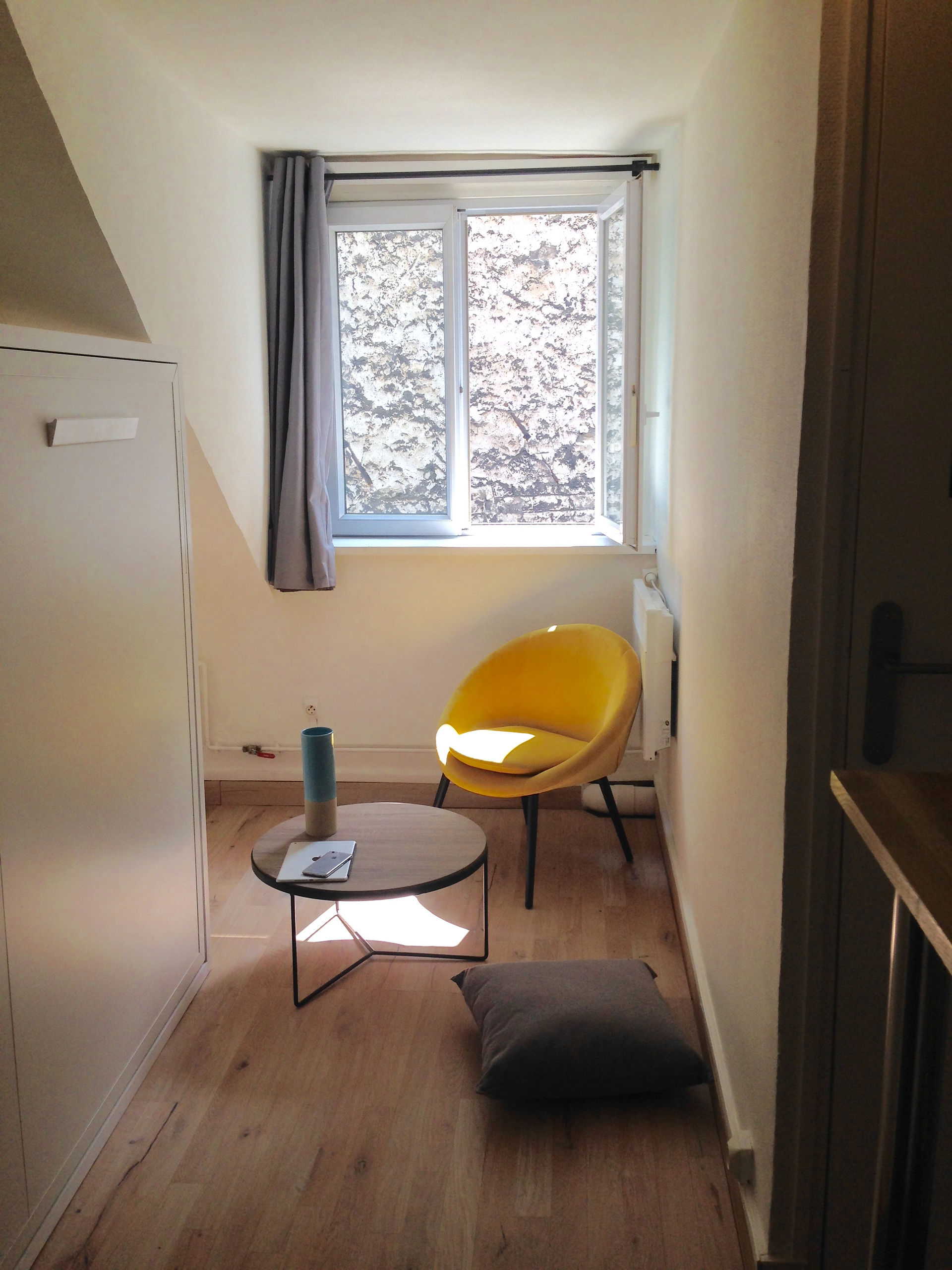 Pièce de vie du studio rue de Cléry, avec fauteuil jaune, table basse et lit rabattable replié.
