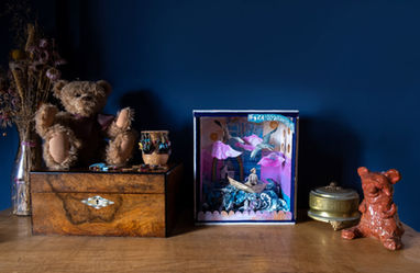 Image of side-table with teddy bear sat on suitcase, trinkets, box 3D artwork with girl sailing in a boat on the sea and whimsical pink clouds
