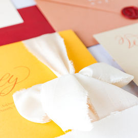 A detailed shot of a wedding suitefeaturing yellow and brown card, white silk ribbon, red wax seal and red and cinnamon envelope.