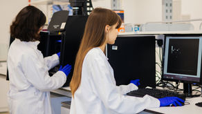 Two researchers in white lab coats and blue gloves working in a laboratory, operating scientific imaging equipment and analyzing data on computer monitors.