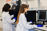 Two researchers in white lab coats and blue gloves working in a laboratory, operating scientific imaging equipment and analyzing data on computer monitors.