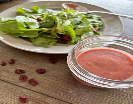 pink cranberry vinaigrette in a small glass bowl. a green salad is in the background with cranberries sprinkled on the table
