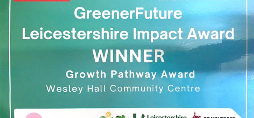 Close-up of the Greener Future Leicestershire Impact Award certificate showing Wesley Hall Community Centre as the Growth Pathway Award Winner, with logos from the University of Leicester, De Montfort University, Leicestershire County Council, National Grid and partners.