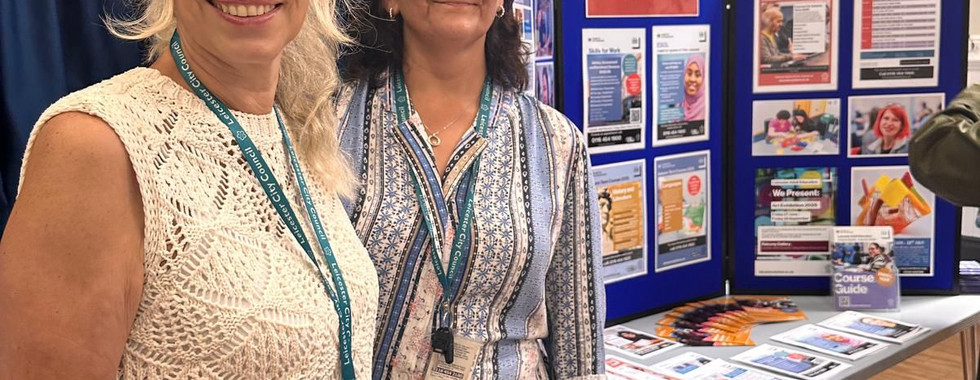 Two Leicester Adult Education representatives smiling at the camera in front of a display board filled with course information and resources at the Wesley Hall Community Centre coffee morning.
