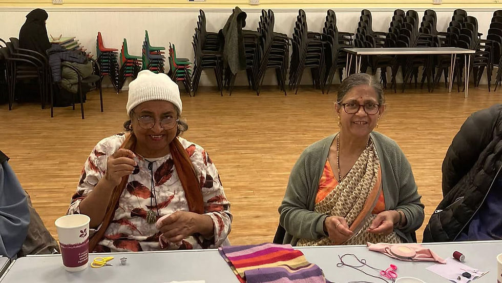 Two women seated at a table, smiling, sewing. One wears a white hat, the other a sari. Stacked chairs in the background, wooden floor.