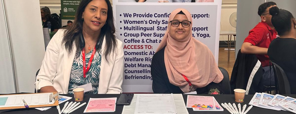 Two representatives from Shama Women’s Centre sitting behind a table covered in leaflets and a logo-branded tablecloth, with a banner promoting confidential support for women.