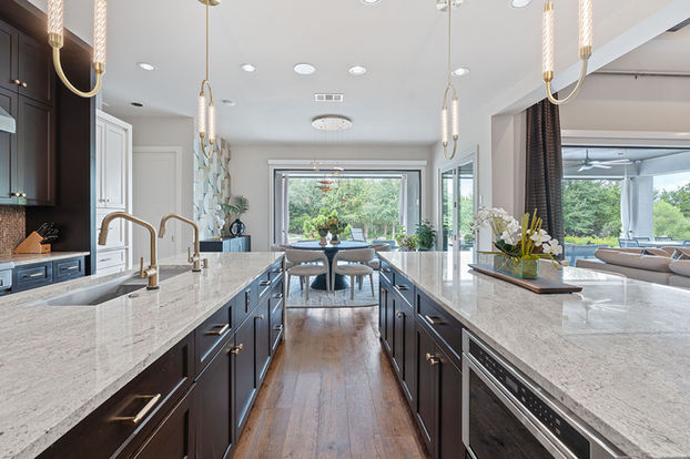 A long perspective view of a gourmet kitchen in a Barton Creek home, showcasing two large islands with waterfall countertops and large windows opening to a patio.