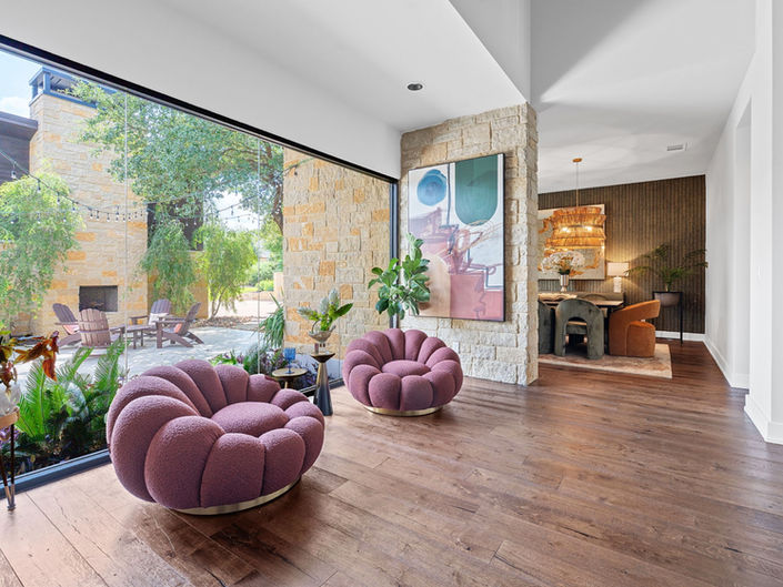 A modern living room in a Barton Creek home with a large glass wall opened to a patio, featuring dark wood floors and two plum colored channel-tufted swivel chairs.