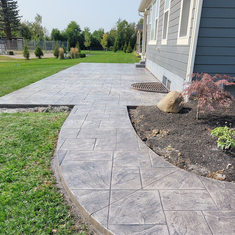 Stamped concrete patio and curved walkway beside a gray house, overlooking a green lawn, trees, and a mulched bed with a rock.