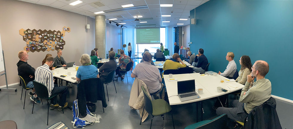 En mötessal med fyra fyrkantiga bord med personer sittande runt och två personer längst fram vid en duk med powerpoint