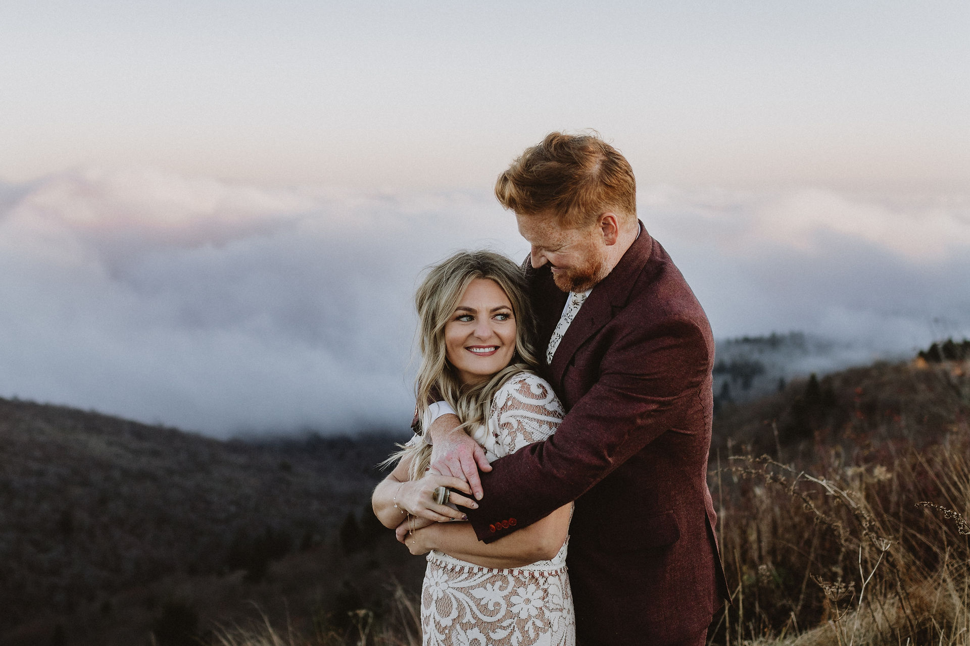 Groom wraps arms around his bride looking down at her, behind them the mountains and low clouds glow blue and pink at sunset