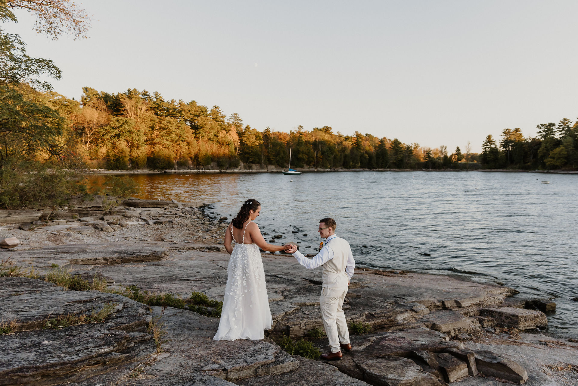 Bride and groom walk along the rocky lakeshore at sunset in the golden hour light
