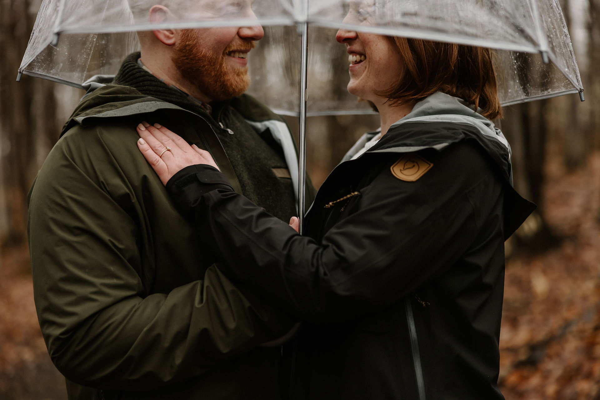 Couple under clear umbrella smiling at each other, both wearing raincoats