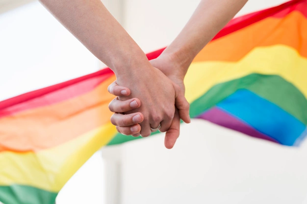 Two people hold hands in front of a rainbow pride flag.