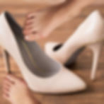 Zwei Damenfüße mit Hallux valgus, im Hintergrund zwei Stöckelschuhe.