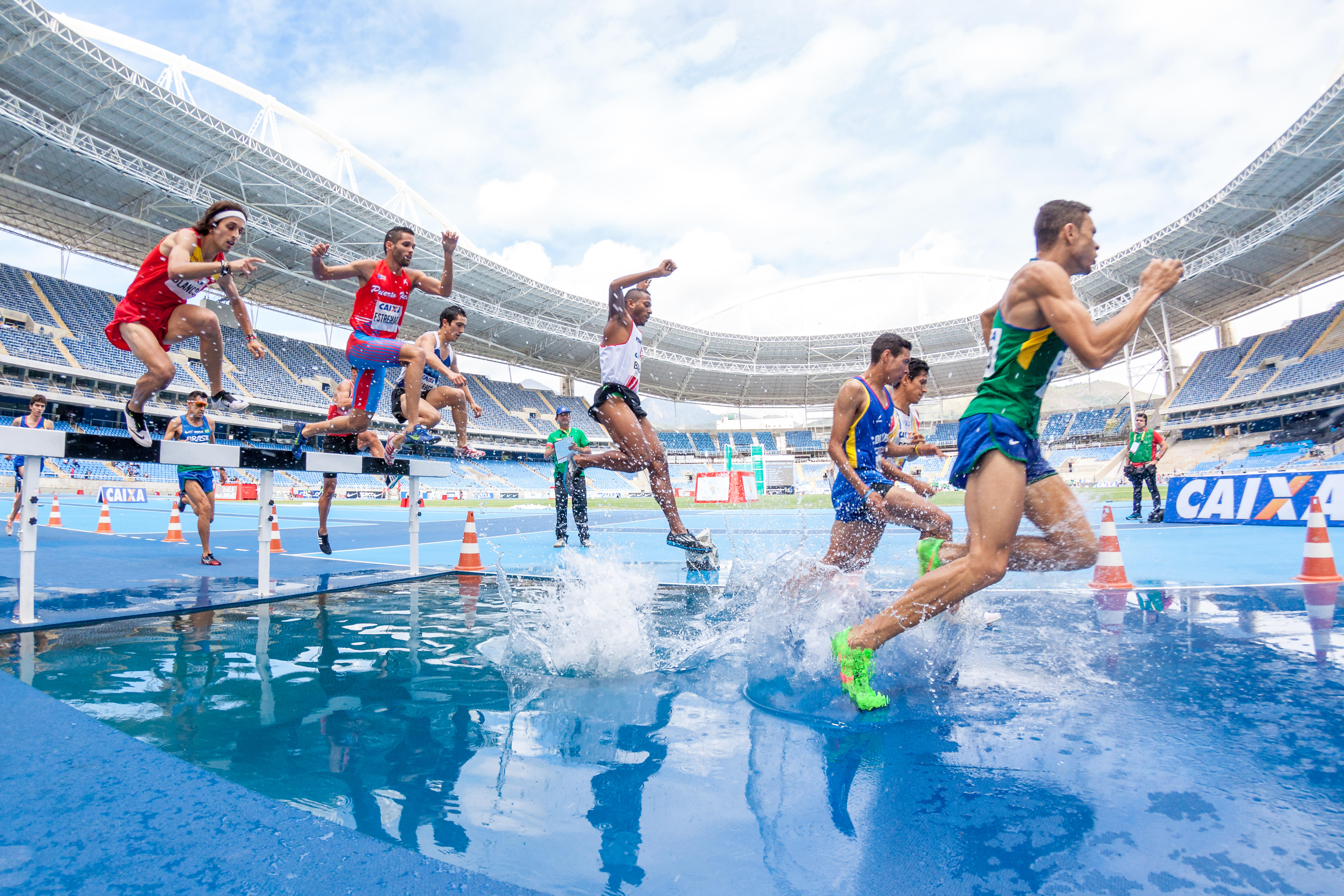 Athletes competing in a steeplechase race in a stadium, jumping over a water obstacle.