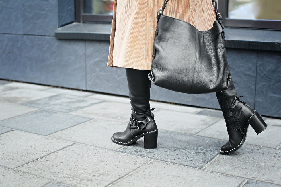 Portrait d'une femme portant des bottine noires, un manteau kaki, tenant un sac à main en cuir noir, vue de face.