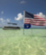 Boat on calm, clear water under blue skies. American and Czech Republic flags on poles, white clouds in the background.