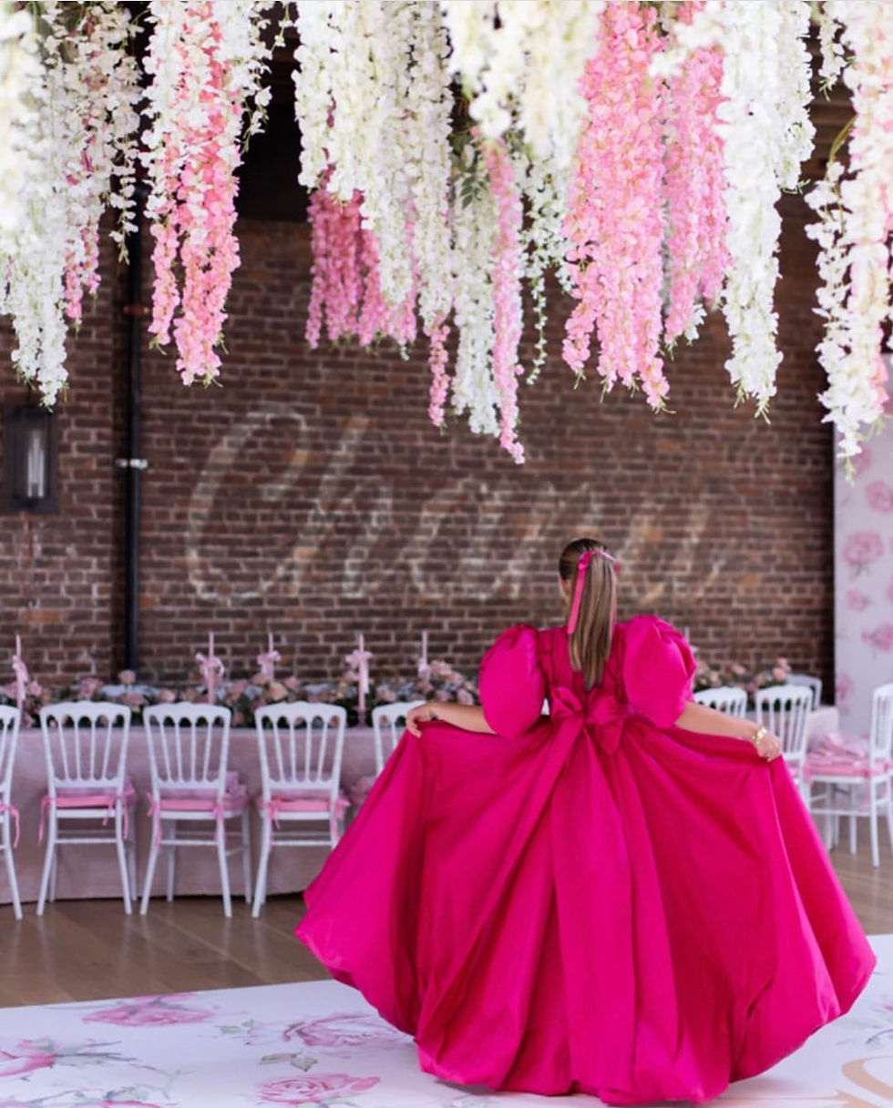 Guest in a bright pink gown walking through floral event space at The Arches BK with hanging white and pink blossoms.
