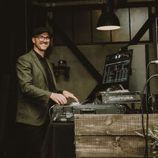 Smiling DJ behind decks in a cosy, rustic space with timber booth and lighting.