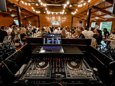 Close view of DJ decks and laptop looking out to a lively wedding reception with string lights.