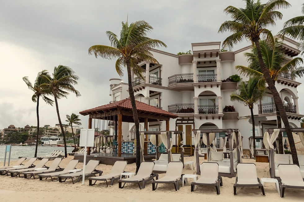 Playa de resort con tumbonas vacías, palmeras y hotel blanco junto al mar bajo cielo nublado, ambiente tranquilo.