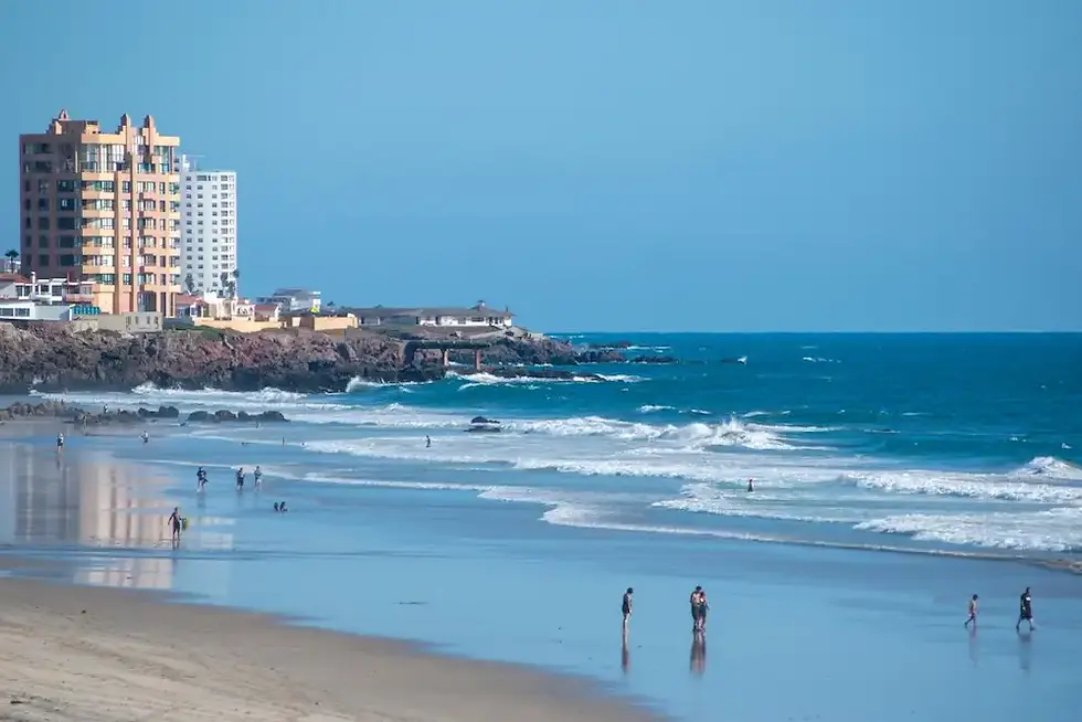 Playa urbana con olas, varias personas caminando en la orilla y edificios altos sobre un acantilado bajo cielo azul.