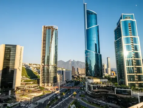 Sunlit city skyline with blue glass skyscrapers, Grand Fiesta Americana tower, mountain backdrop, and busy road below.