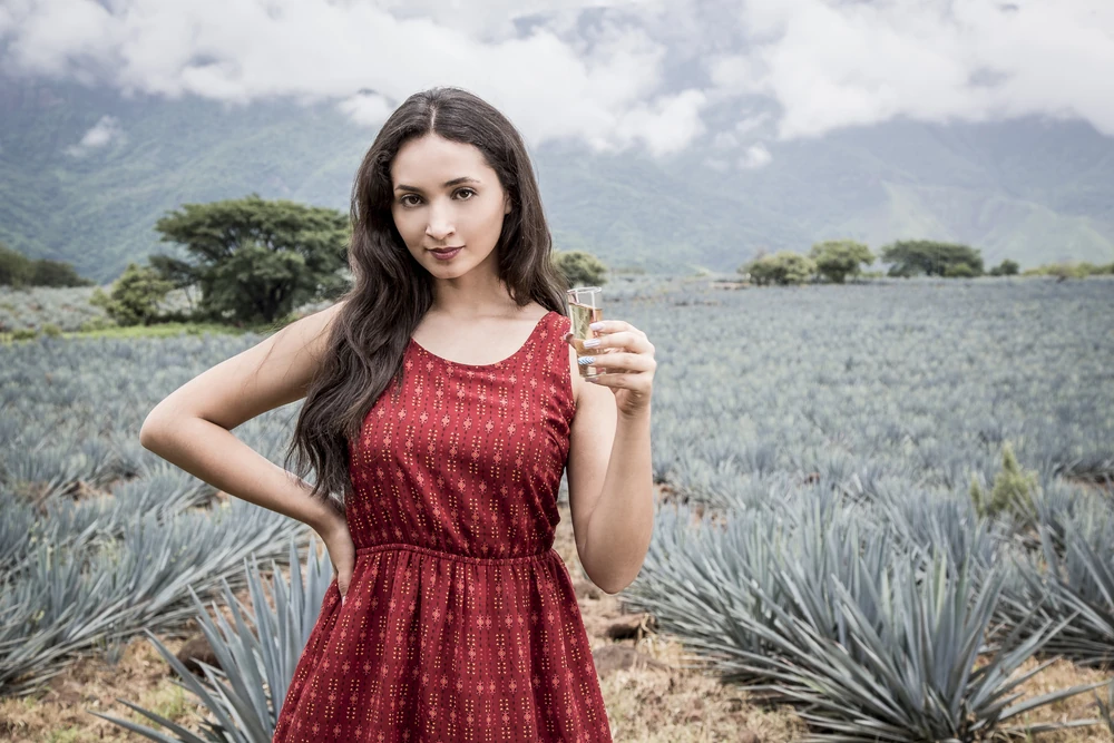 Mujer jóven con un shot de tequila en la mano en medio de plantaciones de agave en Tequila, Jalisco.