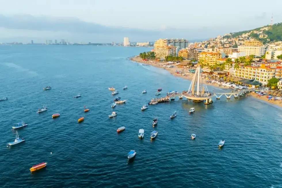 Bahía con muchos botes frente a playa urbana, muelle con velas y colinas llenas de edificios bajo cielo claro.