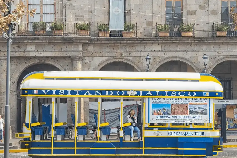 Blue-and-yellow open trolley tour bus labeled VISITANDO JALISCO and GUADALAJARA TOUR stops by a stone arcade, woman seated aboard.