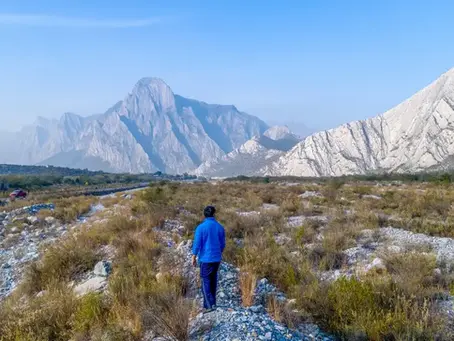 Persona con chaqueta azul camina por terreno rocoso y árido hacia montañas blancas bajo cielo despejado.