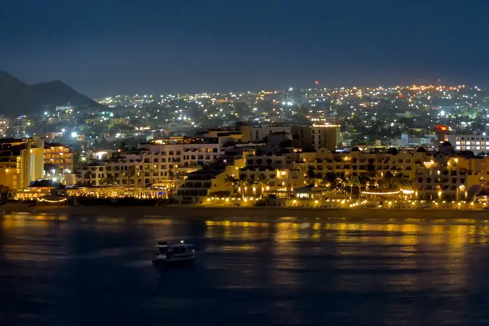 Nighttime waterfront city with glowing resort buildings, a small boat on calm water, and hills lit in the distance.