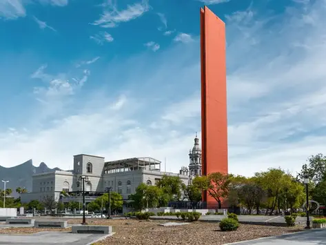 Gran escultura rectangular naranja en una plaza urbana, con edificio histórico, montañas y cielo azul al fondo.