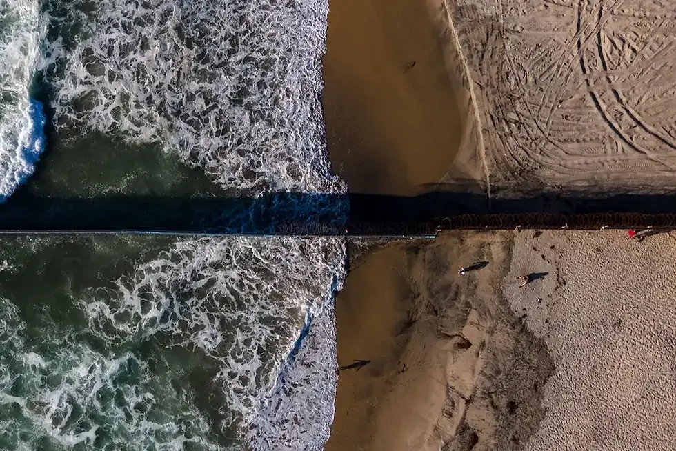 Vista aérea de una playa con olas rompiendo junto a un muelle; dos personas caminan sobre la arena.