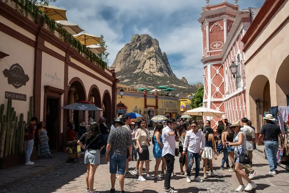 Turistas caminan por una calle colonial con terrazas y letreros, bajo un gran cerro rocoso en un día soleado.