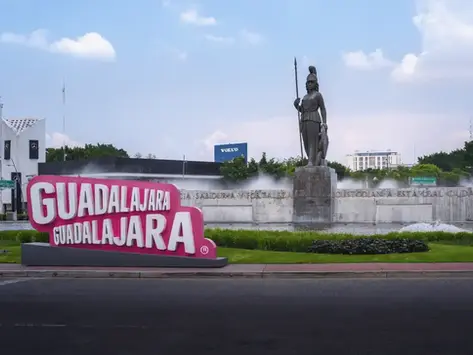 Bright pink Guadalajara sign in front of a fountain and bronze statue, with cloudy sky and city buildings in the background