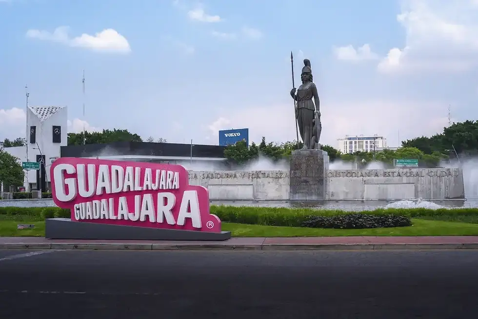 Bright pink Guadalajara sign in front of a fountain and bronze statue, with cloudy sky and city buildings in the background