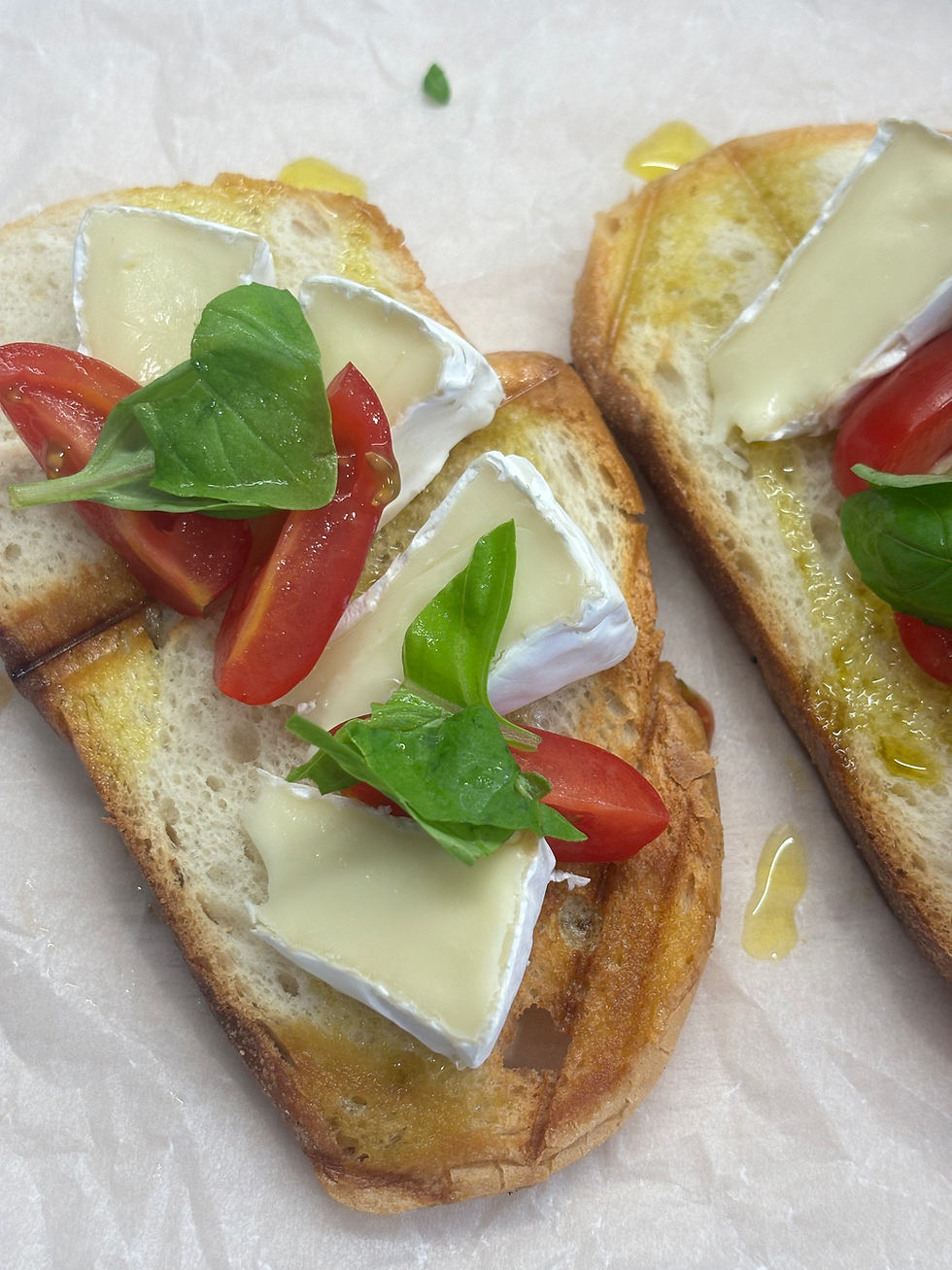 Toasted bread slices topped with brie, cherry tomatoes, basil, and olive oil on parchment, a fresh appetizer close-up