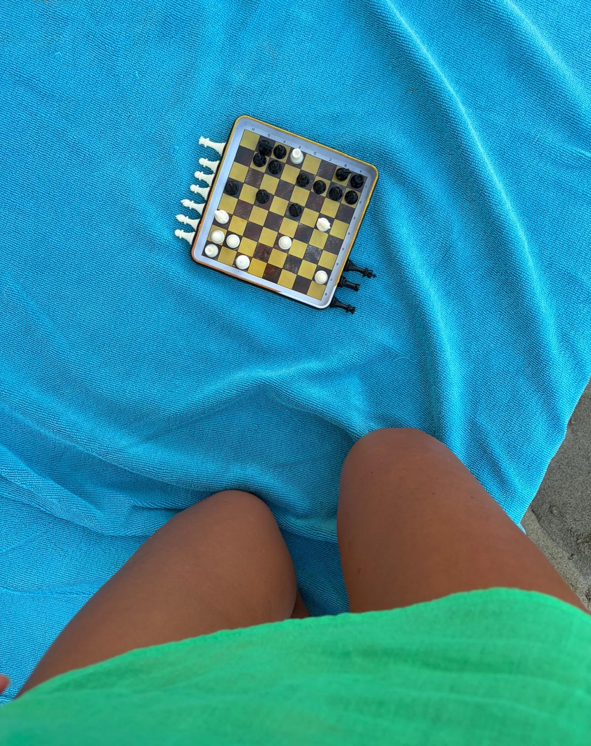 Small chessboard with numbered edges on a bright blue towel between a person’s legs, near beach sand.